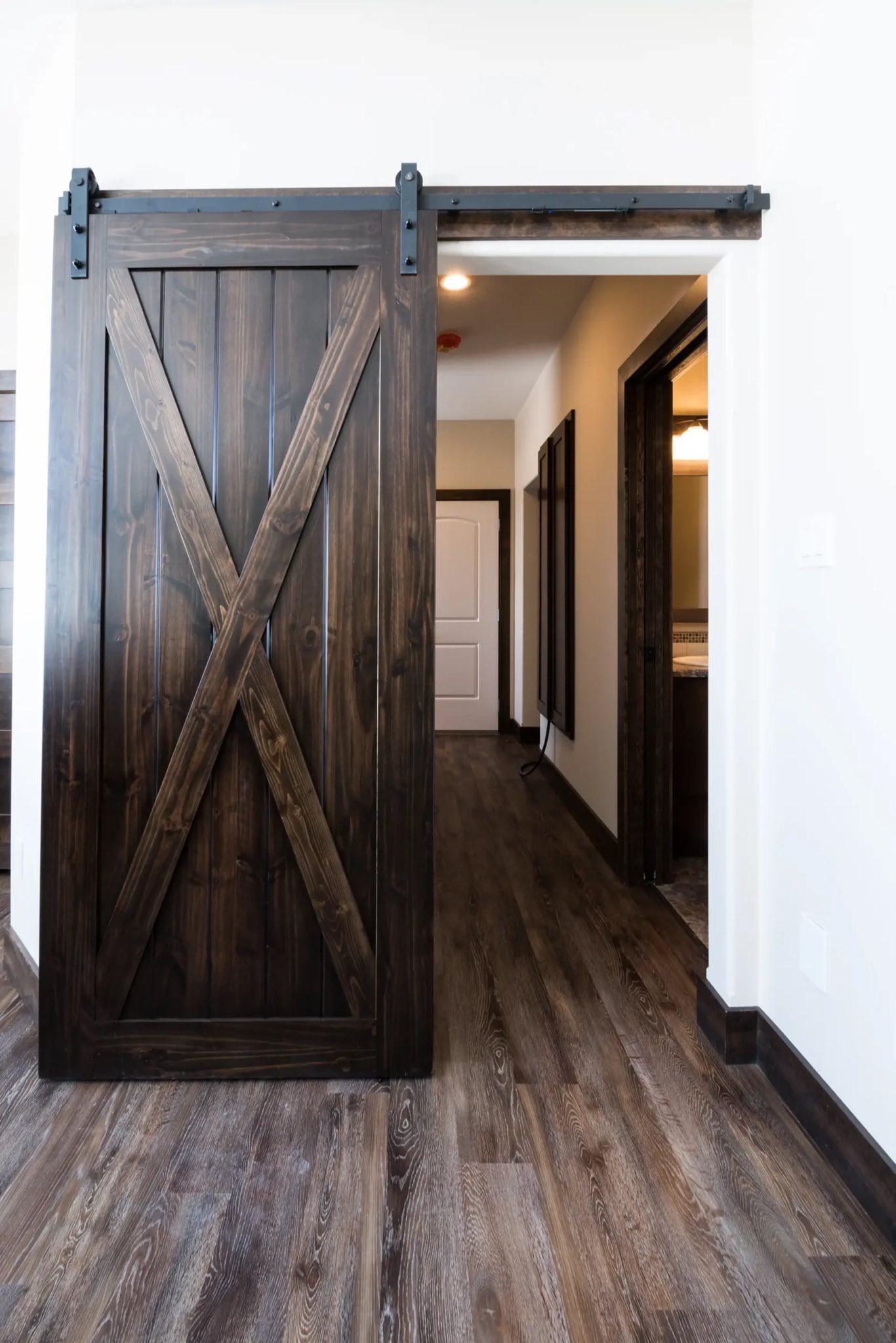 Dark wooden barn door with a prominent X design slides open to reveal a hallway with smooth, dark wood flooring and warm-toned walls, conveying a rustic and cozy atmosphere.