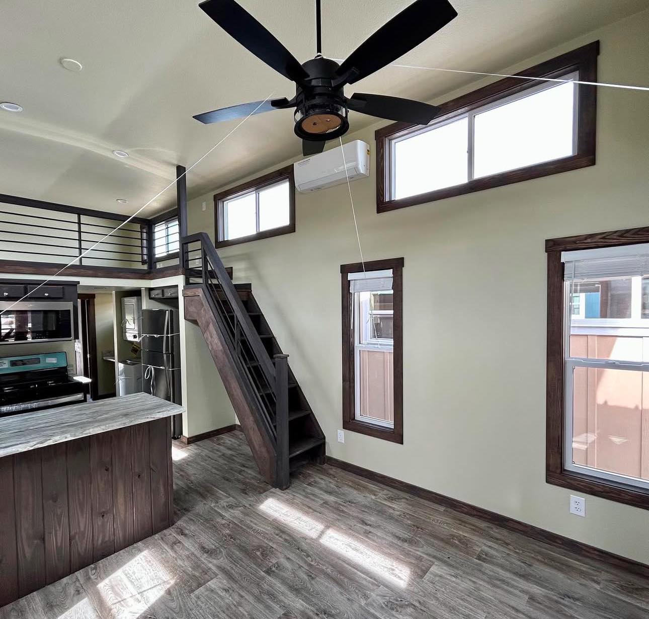 A modern tiny house interior with high ceilings, featuring a loft accessible by wooden stairs, large windows, a ceiling fan, and a kitchen area with appliances.
