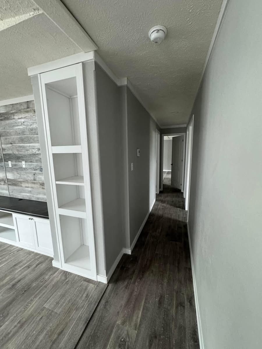 Narrow hallway with gray walls and dark wood flooring. Shelving built into the left wall, leading to an open door at the end. Bright and modern.