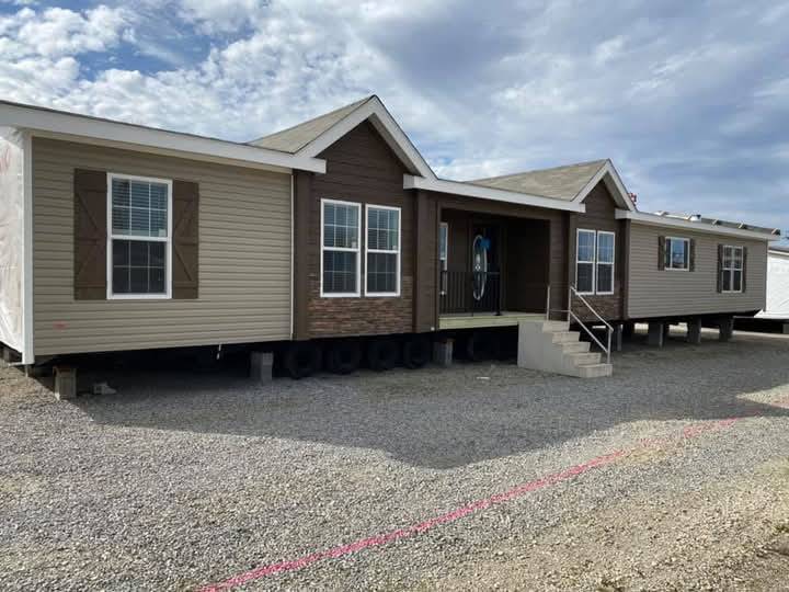 Manufactured home with beige and brown siding, elevated on blocks, against a cloudy sky. Steps lead to the porch, creating a welcoming entrance.