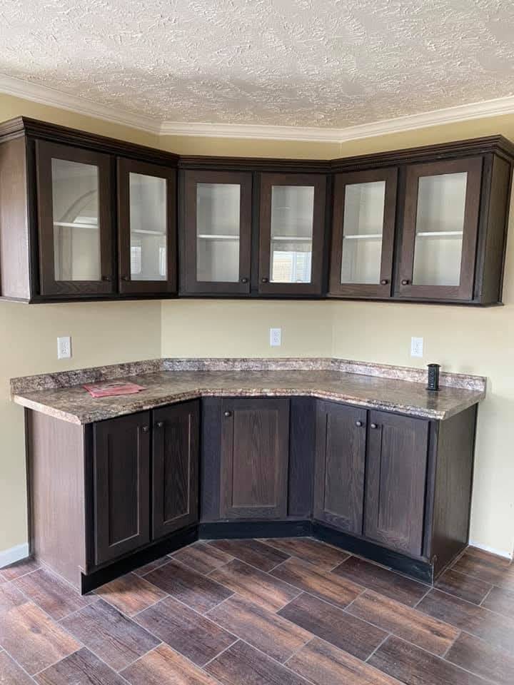 Dark wooden corner kitchen cabinets with glass door fronts and brown countertops. Textured ceiling and tiled floor create a cozy, rustic feel.