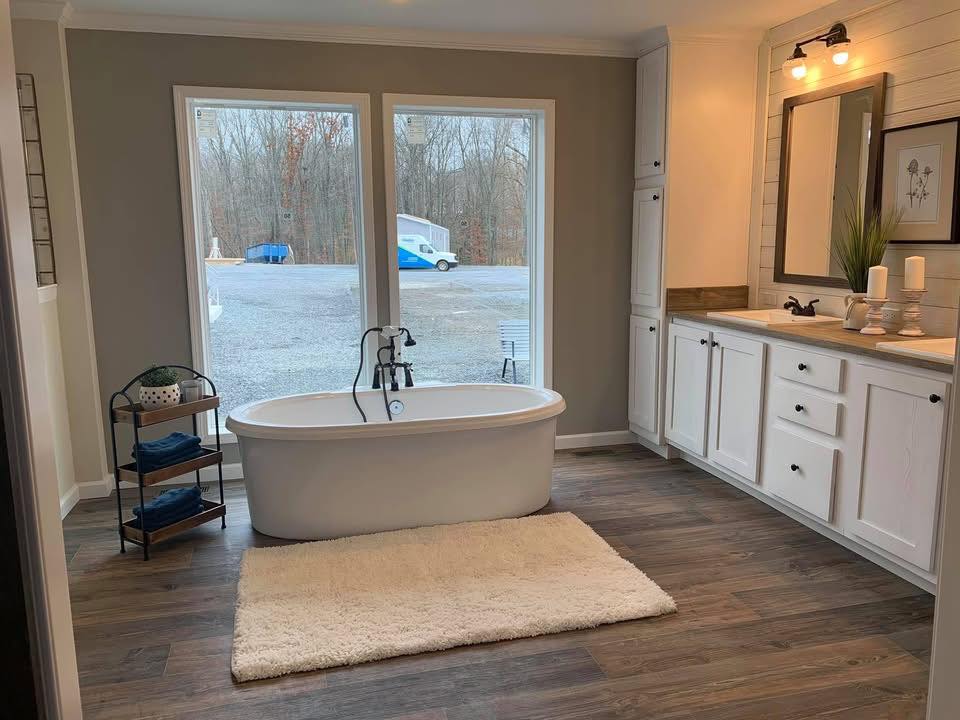 A serene bathroom with a freestanding tub on a beige rug, flanked by large windows showing a wooded view. White cabinets, dark fixtures, and wood accents.