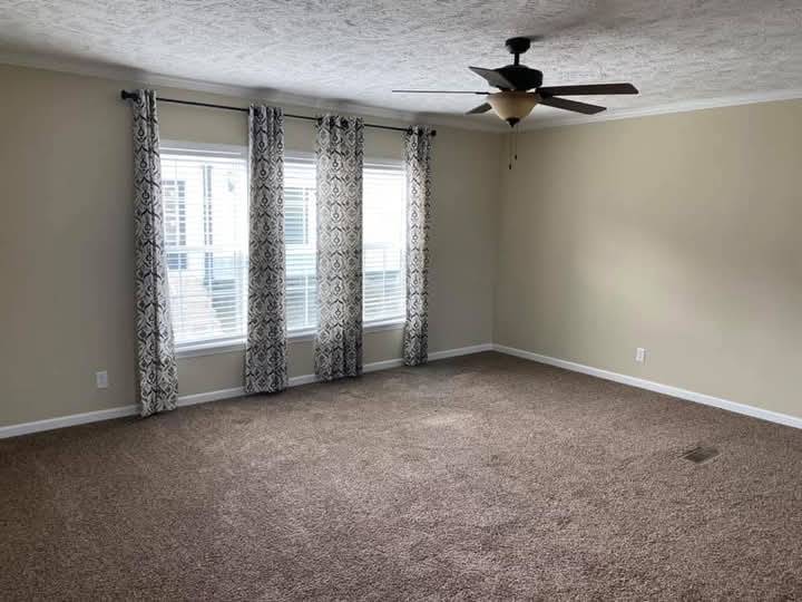 A spacious, empty room with beige walls and brown carpet. Large windows with patterned curtains and a ceiling fan create a calm, inviting atmosphere.