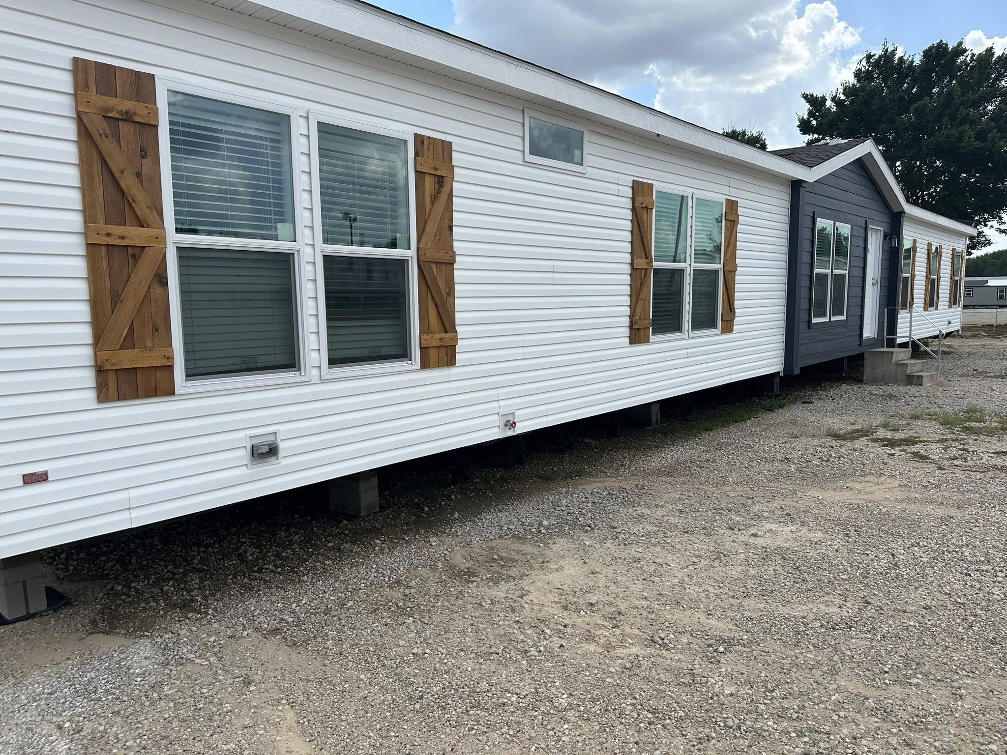 Large white mobile home with wooden shutters and a dark gray section, set on a gravel lot. It is positioned under a partly cloudy sky.