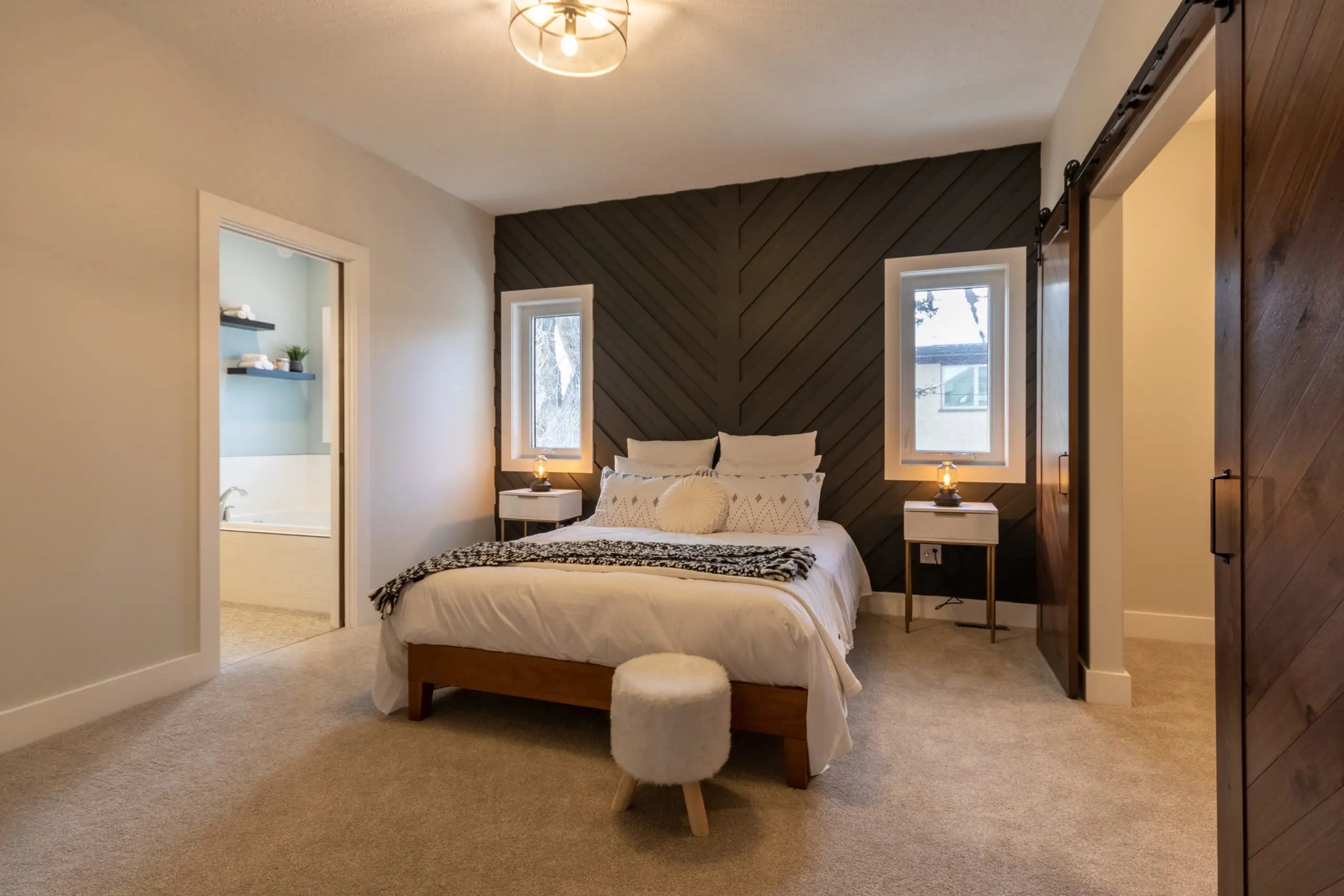 A modern bedroom with a cozy, neutral tone. Features a bed with white linens, chevron-patterned dark accent wall, two windows, two side tables, and warm lighting.