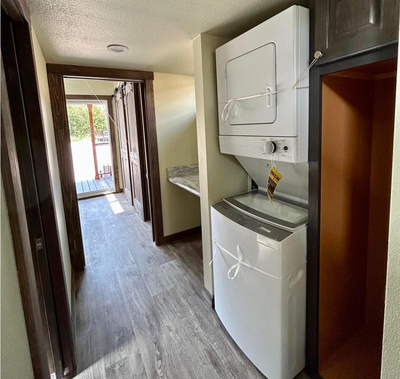 Narrow hallway with wood floors leading to a sunlit porch. On the right, a white stacked washer and dryer is tied with a cord. Cozy and functional.