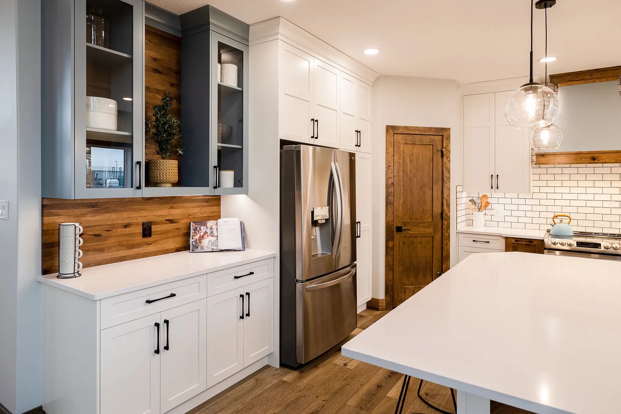 Modern kitchen with white cabinets, stainless steel fridge, and wooden accents. A large island with pendant lights adds a sleek, inviting touch.