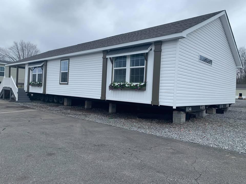 A white manufactured home sits elevated on blocks in an outdoor lot. It features large windows with flower boxes, under an overcast sky, creating a tranquil atmosphere.