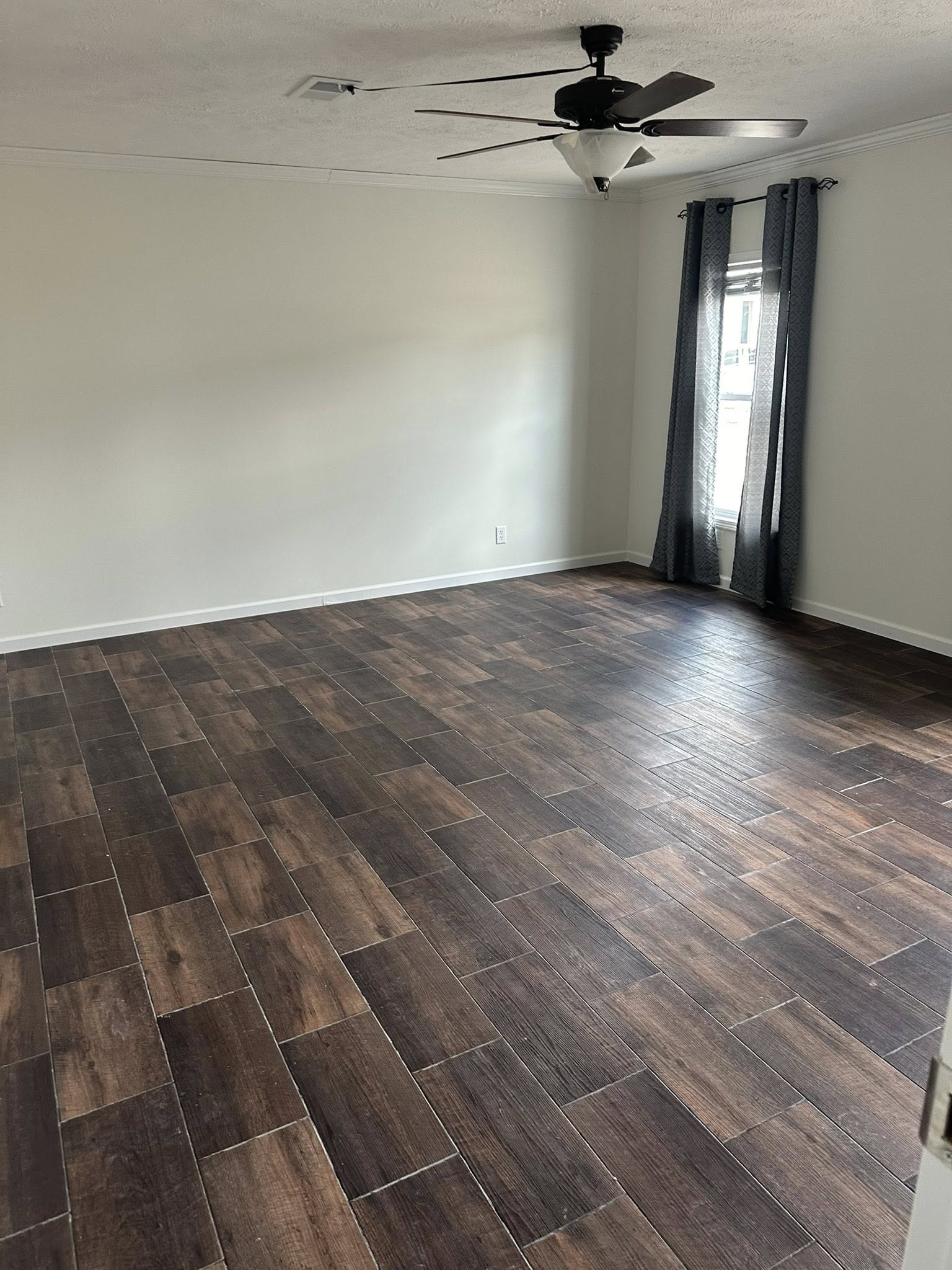 Empty room with dark brown wood flooring, white walls, and a ceiling fan. A window on the right wall has gray curtains. The room feels spacious and clean.