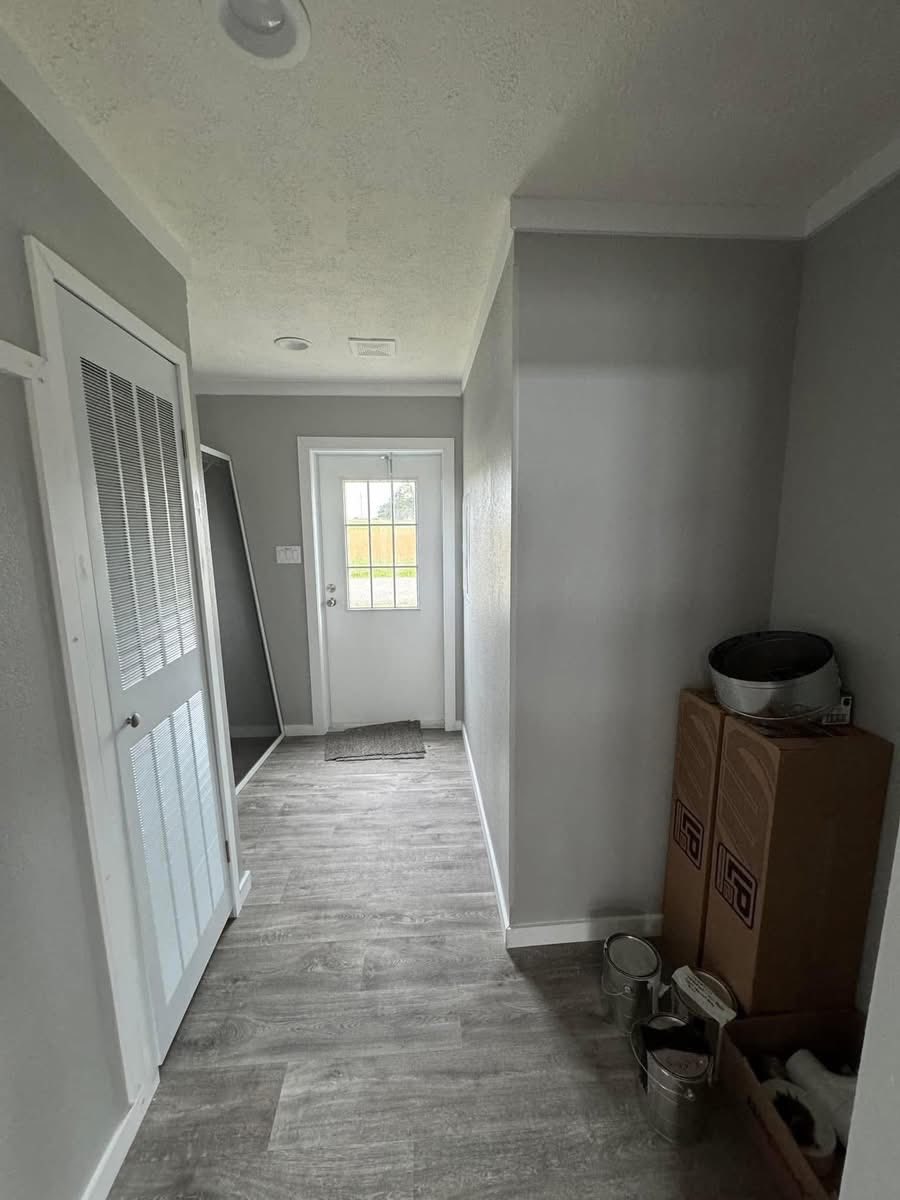 A hallway with gray walls and light wooden floor leads to a door with a window. On the right, boxes, paint cans, and a bucket are stacked. The scene is tidy and minimalistic.