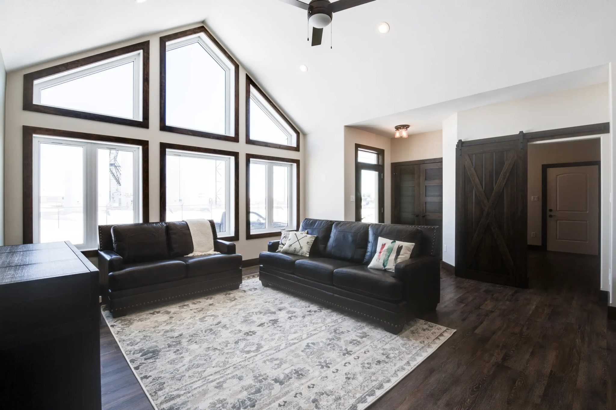 Airy living room with high ceilings, prominent triangular windows, dark leather sofas on a patterned rug, and rustic barn door on wooden floors.