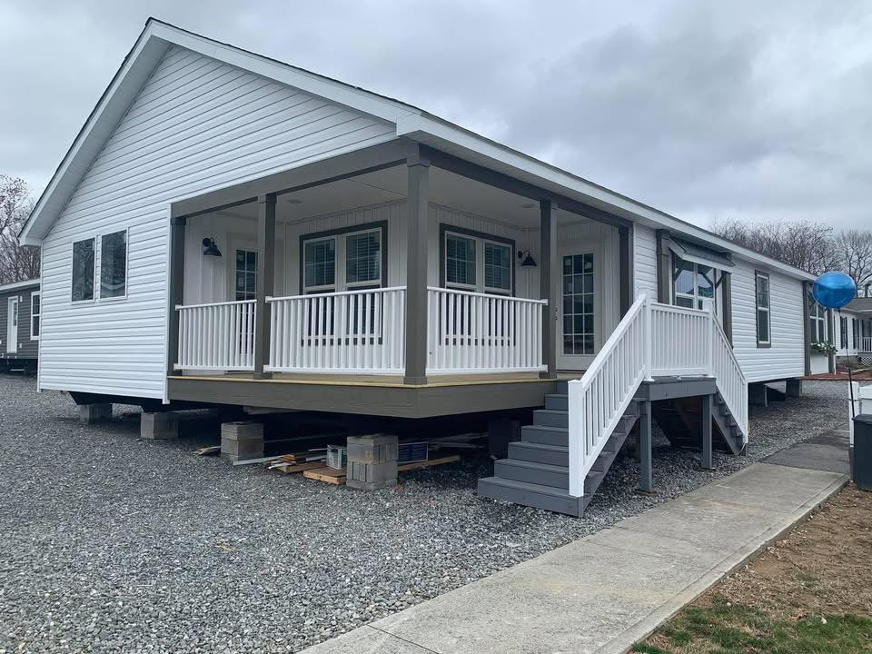 Single-story white modular home with a covered porch, wooden steps, and a gravel foundation. Overcast sky creates a neutral, calm atmosphere.