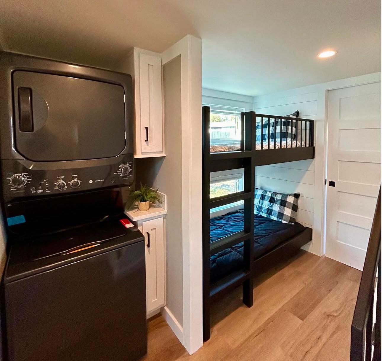 Compact room with stacked black washer-dryer and bunk beds. The top bed has plaid bedding. Warm hardwood floor, white walls, and soft lighting. Cozy and efficient.
