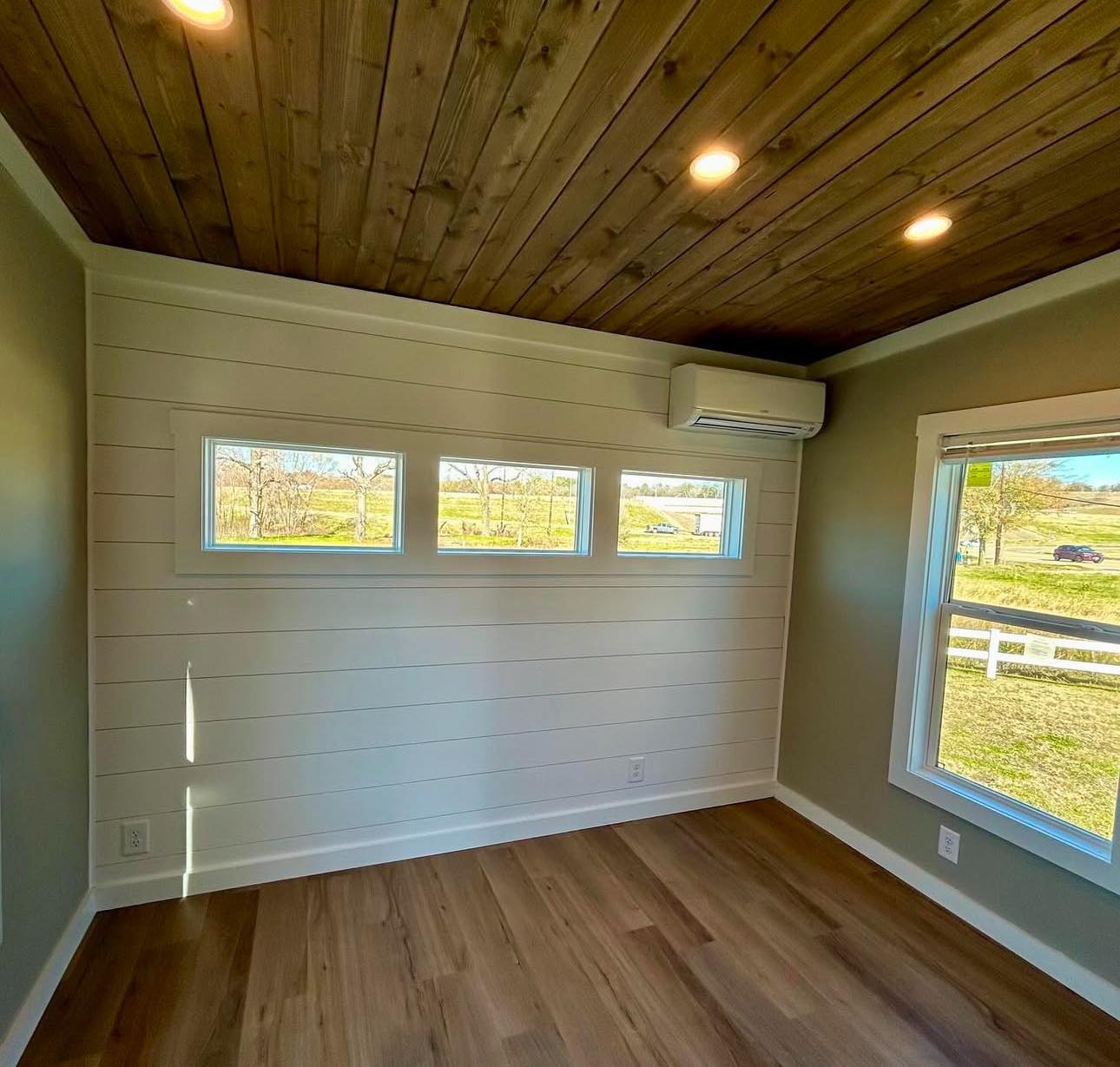 Small room with wood ceiling, recessed lights, white shiplap walls, and large windows showcasing a scenic outdoor view. Air conditioner mounted high.