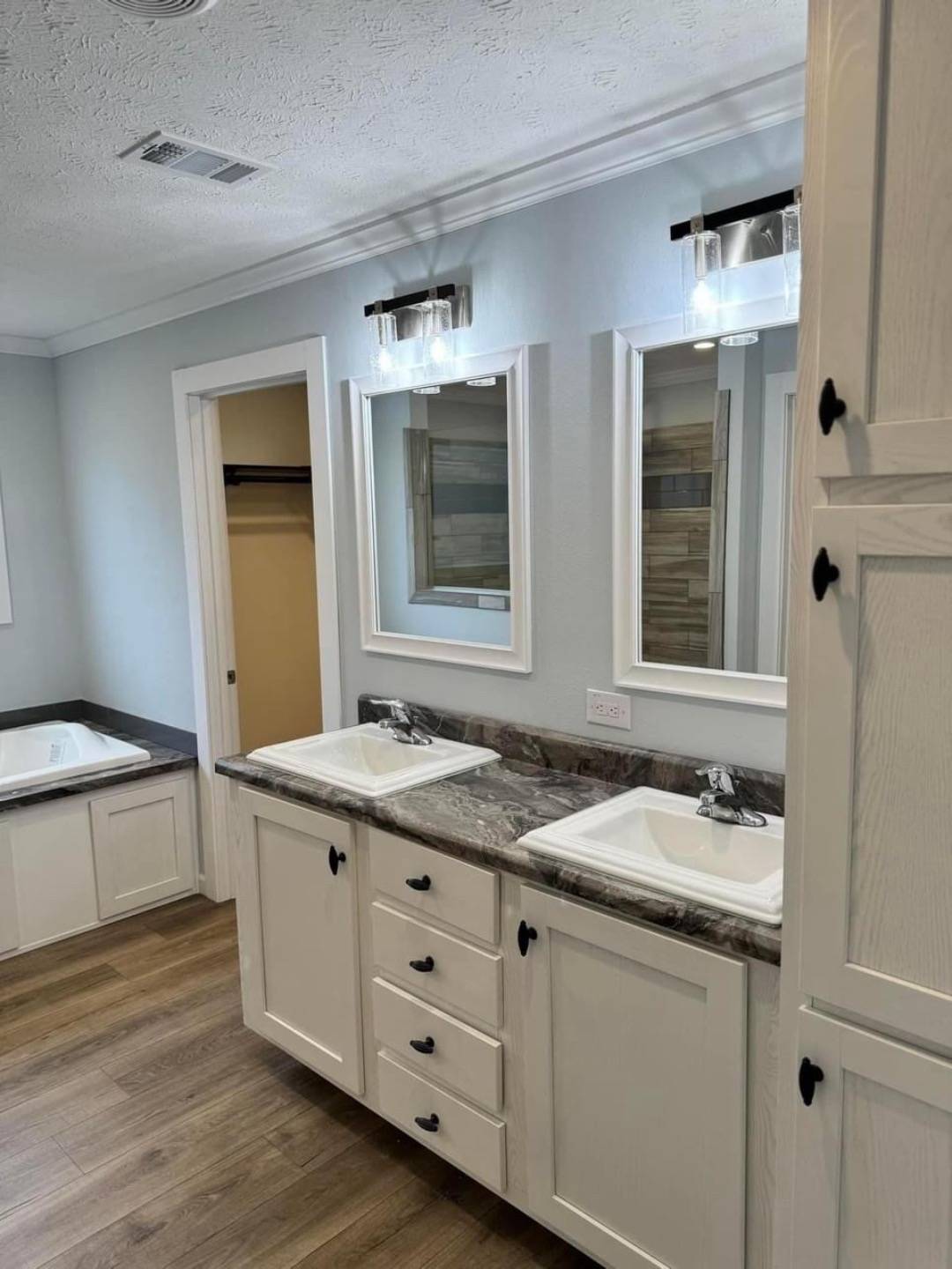 Modern bathroom with light blue walls, dual sinks on a marble countertop, wooden cabinetry, large mirrors, and bright overhead lights; a cozy, clean ambiance.