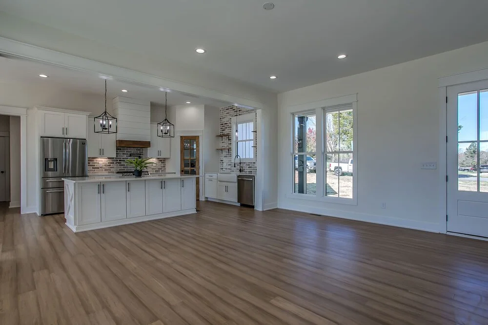 Spacious modern kitchen with light wood floors, white cabinets, and stainless steel appliances. Large windows and pendant lights create an airy feel.