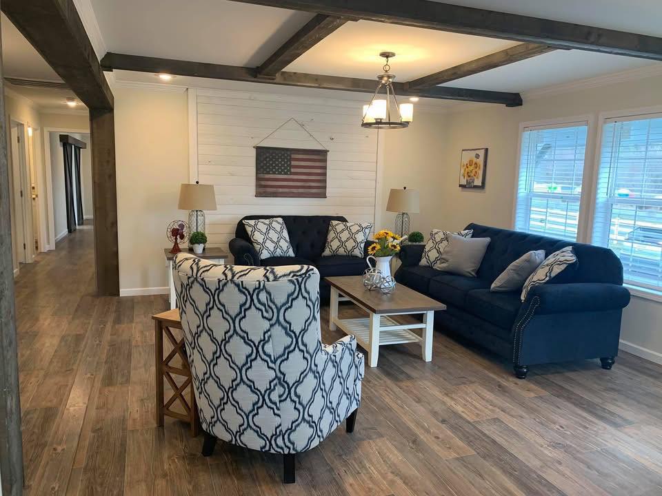 Cozy living room with wooden floor, navy sofas, and patterned armchair. American flag art on wall, sunflower decor, and soft lighting create a warm ambiance.