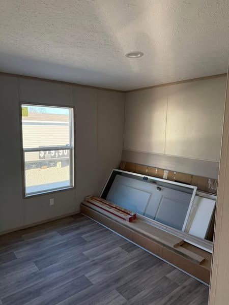 A small, unfurnished room with gray laminate flooring and neutral walls. A window on the left lets in light, illuminating unassembled furniture against the wall.