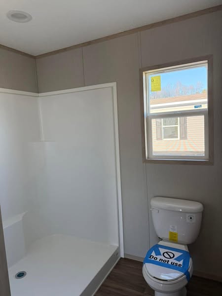 A simple bathroom with a shower, unopened toilet labeled "Do not use," wood flooring, gray walls, and a window showing a neighboring building.