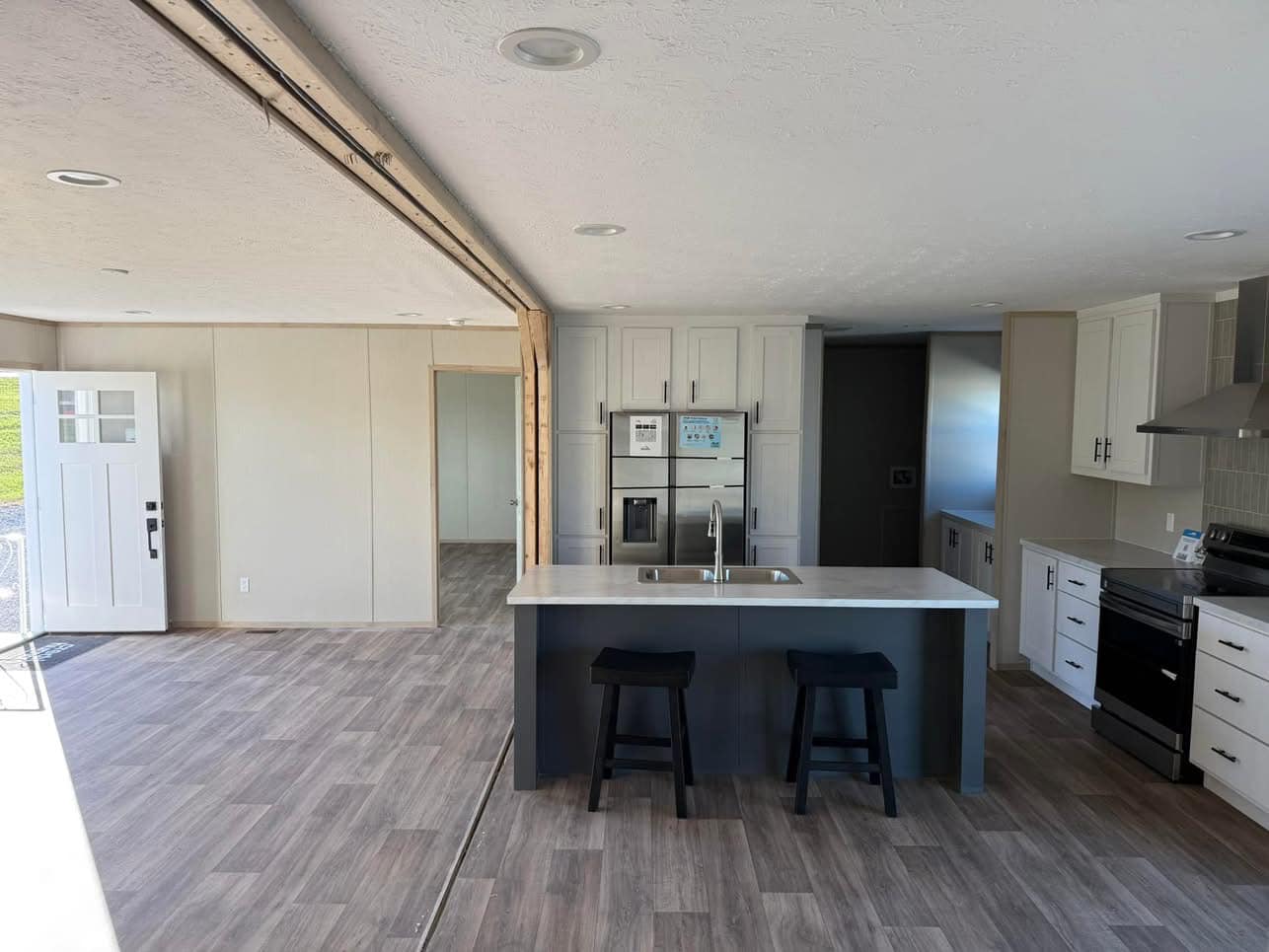 A modern kitchen with light wood flooring, white cabinets, and a central island with two black stools. The room is brightly lit and spacious.