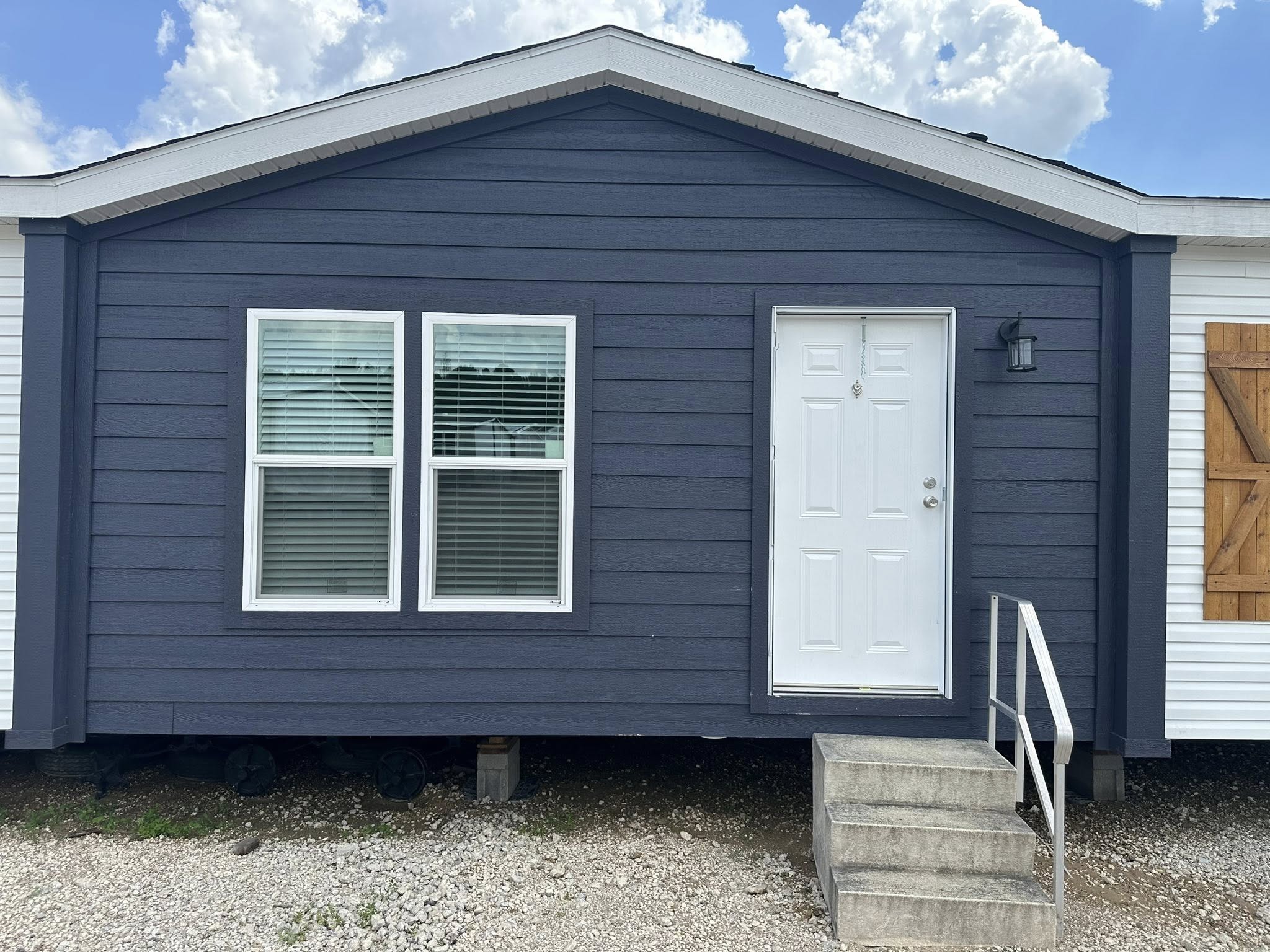 Gray mobile home with a white door, double windows, and a small staircase. The sky is bright with scattered clouds, creating a calm atmosphere.