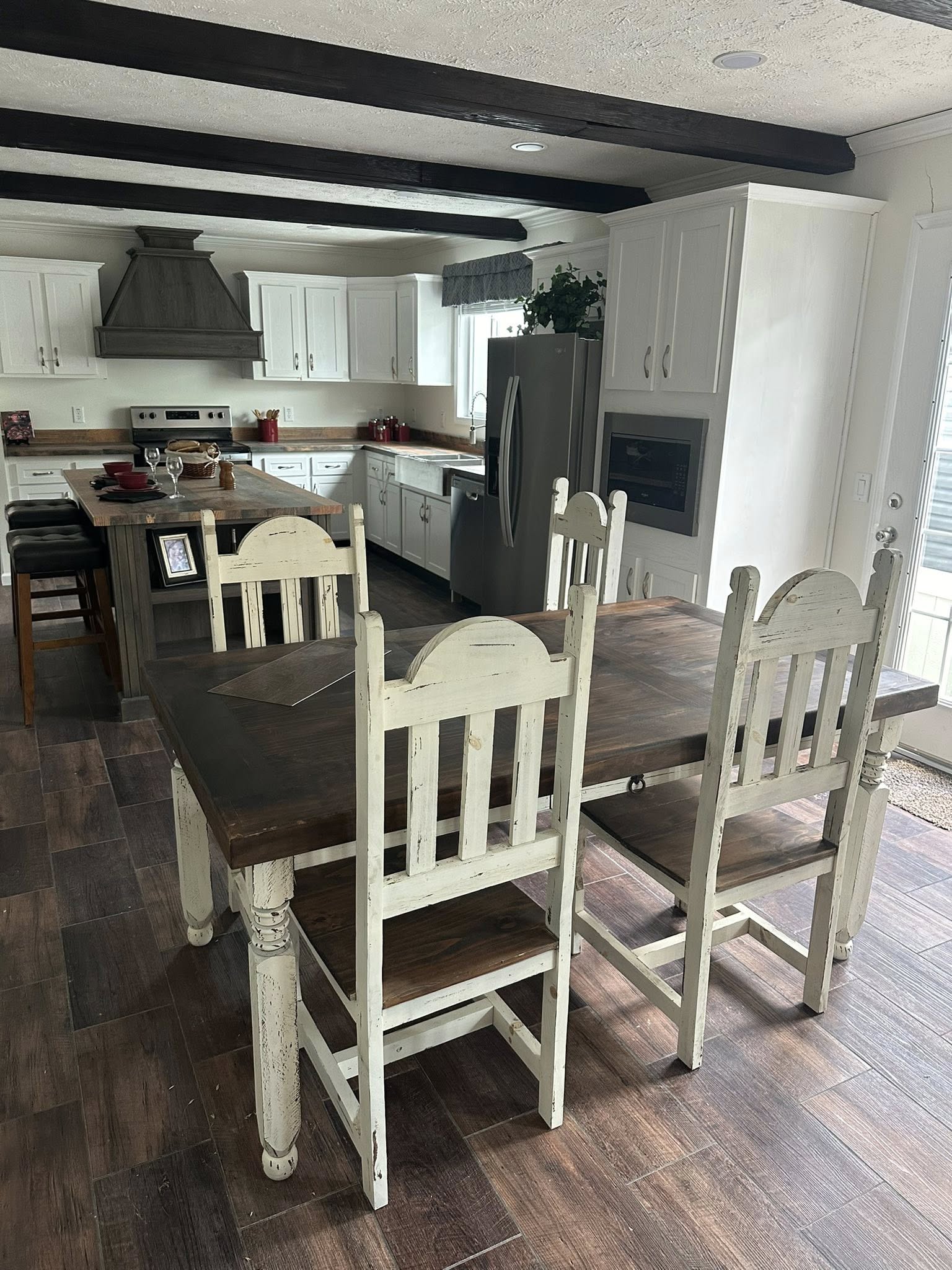 Rustic kitchen with dark wooden floors, white cabinets, and exposed beams. A wooden dining table with distressed chairs creates a cozy, welcoming atmosphere.