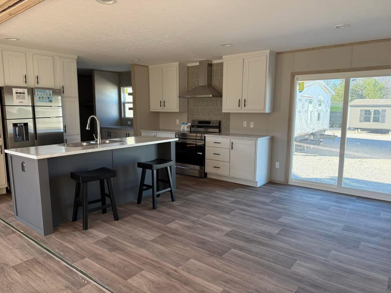 Modern kitchen with light wood flooring, white cabinets, and stainless steel appliances. Island with two black stools, large window on the right. Bright and spacious.