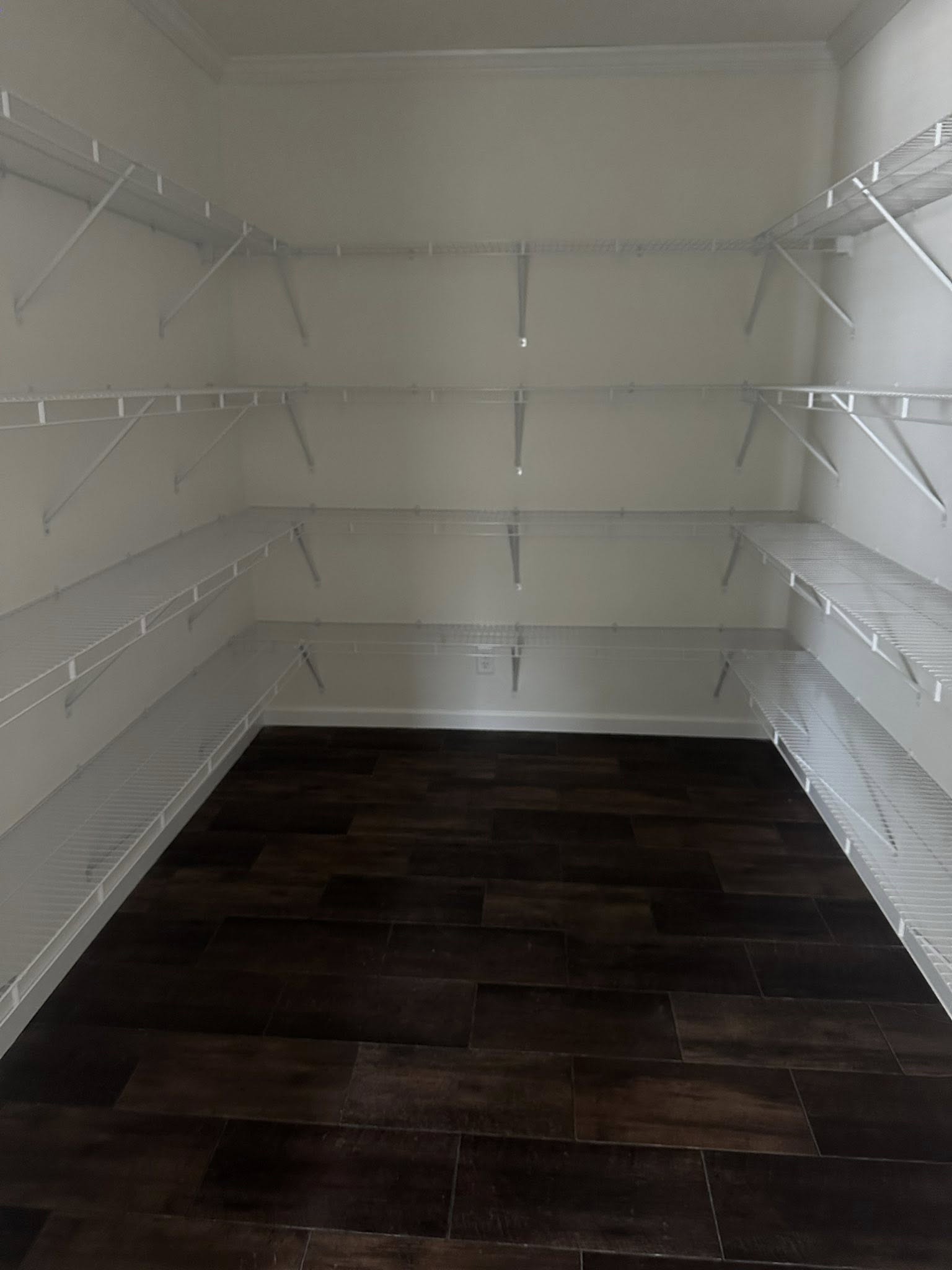 Empty walk-in pantry with white wire shelving on three walls and dark wood flooring. The space is well-lit, appearing clean and organized.