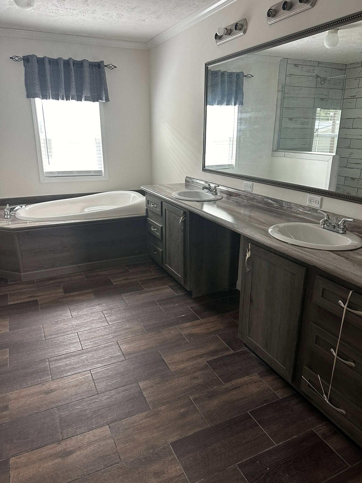 Spacious bathroom with dual sinks on a dark wooden vanity, a large mirror, and a bathtub below a window with blue curtains. Warm, elegant ambiance.