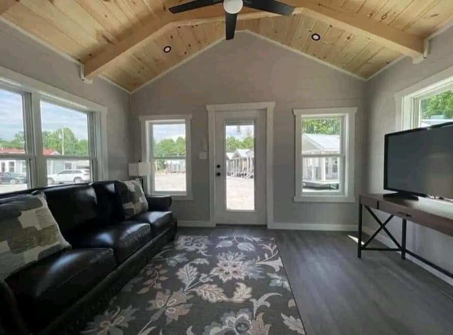 Cozy living room with a vaulted, wood-paneled ceiling, dark leather sofa, floral rug, and large TV on a stand. Sunlight streams through multiple windows.
