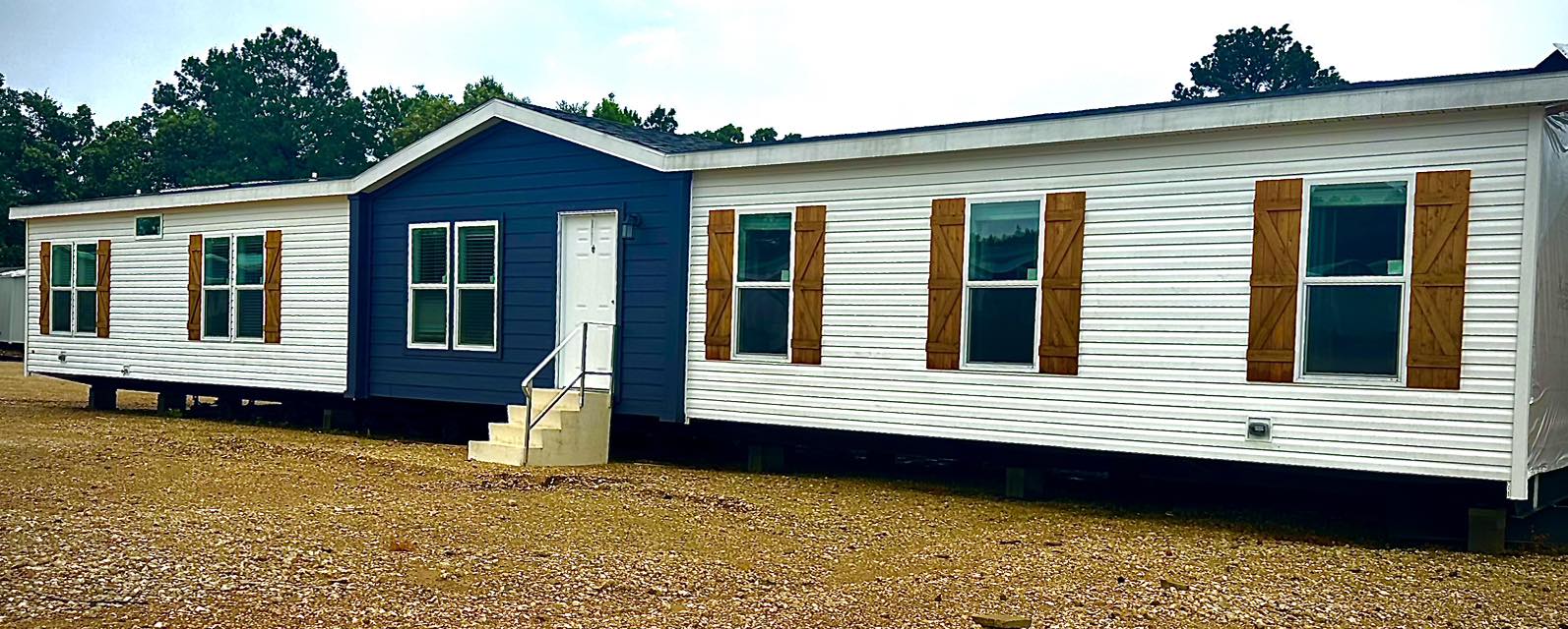 A manufactured home with blue siding and white trims sits on a gravel lot. Large windows with wooden shutters add a rustic touch. Trees are visible in the background, creating a serene ambiance.