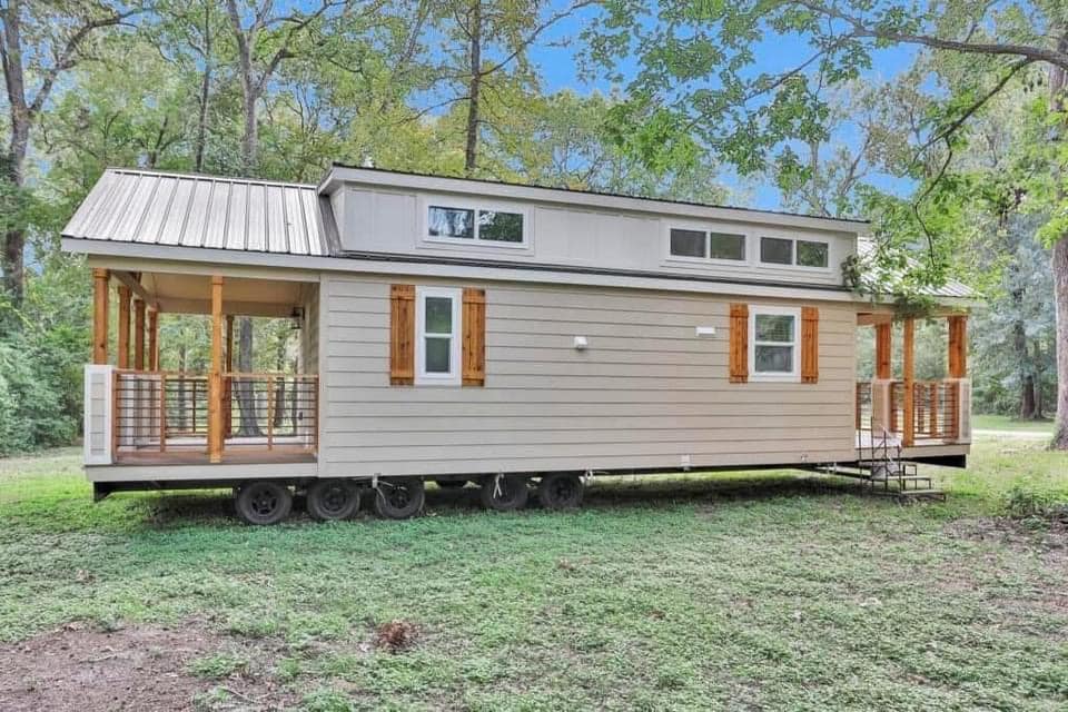 A beige tiny house on wheels with wooden shutters and a metal roof, surrounded by lush trees. The serene setting conveys a peaceful, rustic vibe.