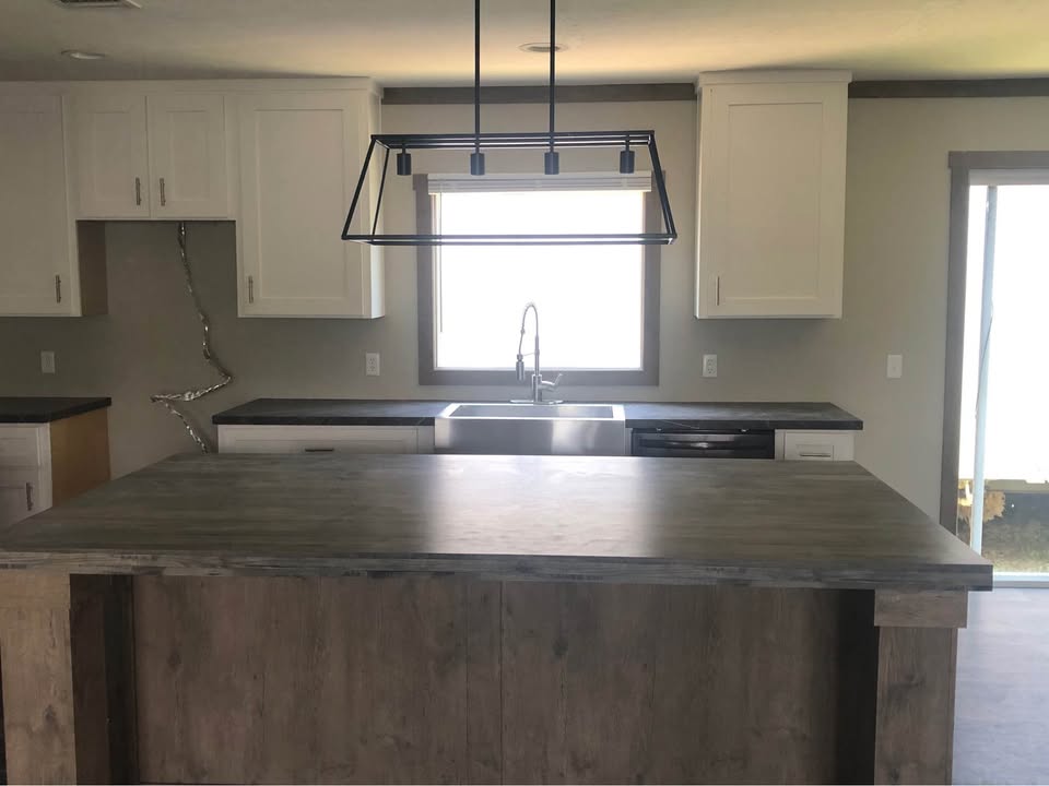 Modern kitchen with a large wooden island in the foreground, white cabinets, and a sleek metal light fixture. Sunlight streams through a window above the sink.