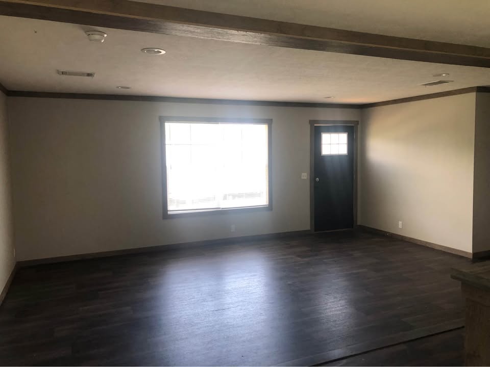 Spacious empty room with dark hardwood floors, beige walls, a large window letting in natural light, and a closed black door, creating a calm ambiance.