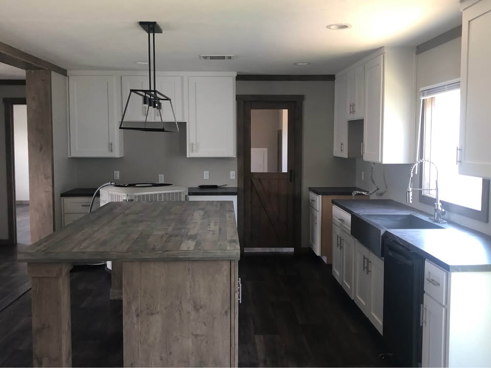 Modern kitchen with a rustic feel, featuring a wooden island, white cabinets, dark countertops, a farmhouse sink, and pendant lighting. Bright and inviting.
