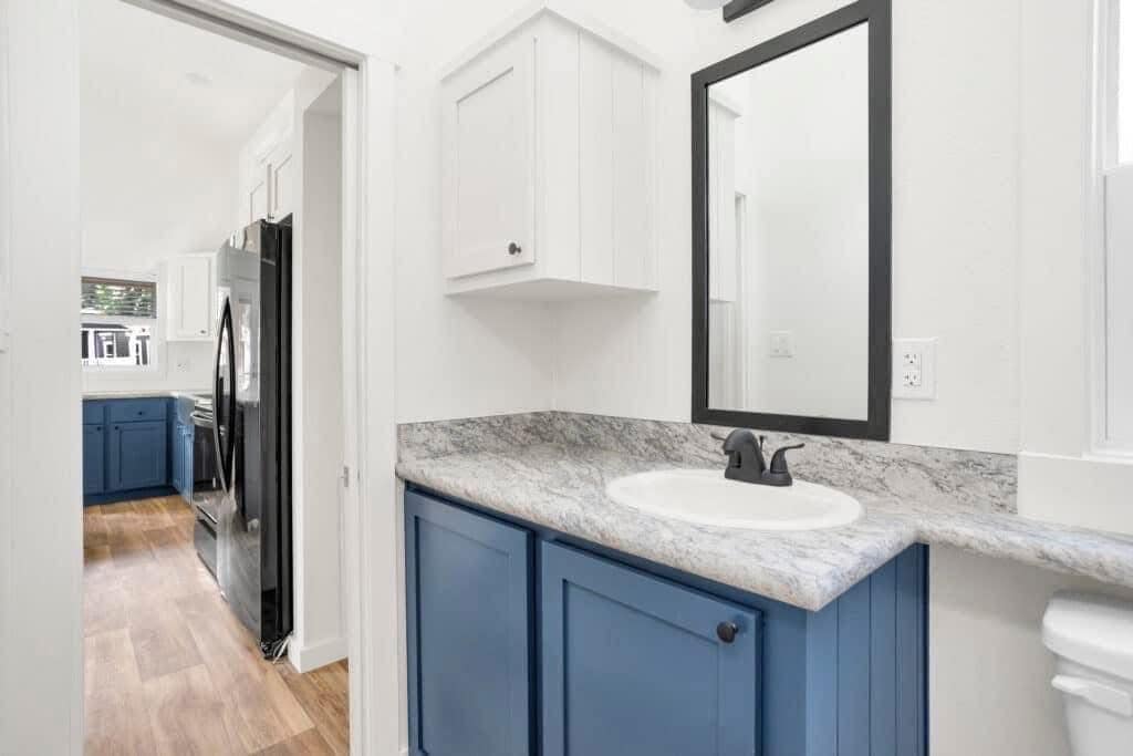 Small bathroom with blue cabinets, a marble countertop, and a black-framed mirror. Adjacent kitchen with wood flooring is visible through the doorway.