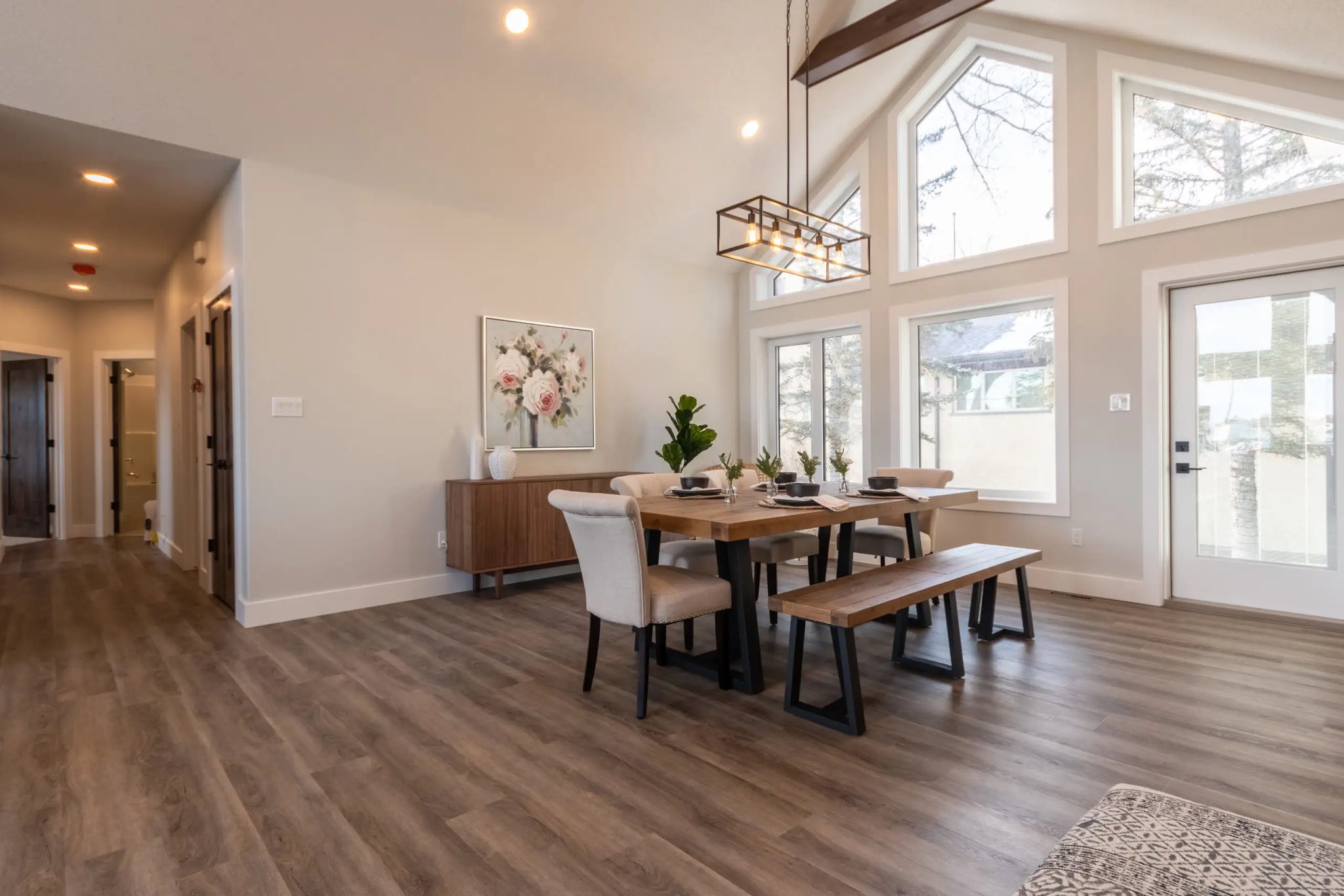 Spacious dining room with large windows, a wooden table set with a bench and chairs, and modern lighting. Warm tones and floral artwork create an inviting atmosphere.