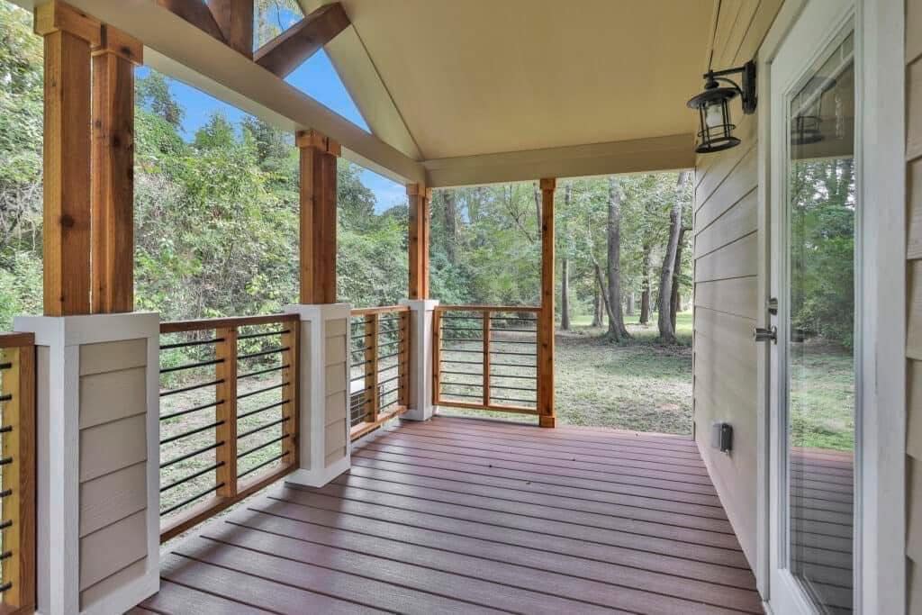 Covered wooden porch with railing overlooks a grassy area surrounded by trees. The porch has large beams, a slanted roof, and a glass door.