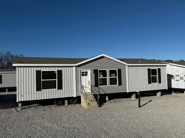 A gray modular home with a gabled roof and white door sits on a gravel lot. It features black shutters, three steps leading to the entrance, under a clear blue sky.