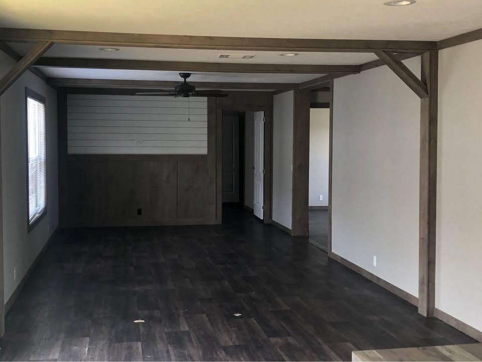 Empty room with dark wood floors, light beige walls, and wooden beams. A ceiling fan hangs above, and doors are visible in the back. Natural light enters from a window on the left, creating a calm atmosphere.