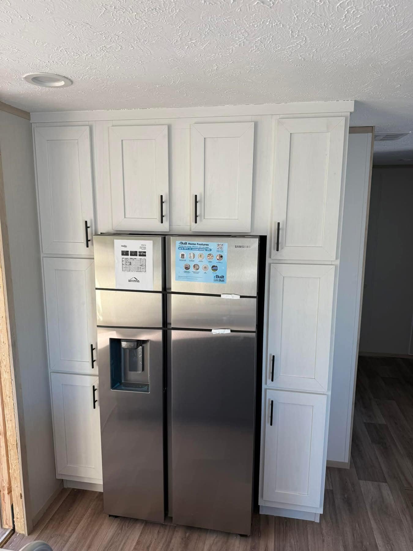 Stainless steel refrigerator with water dispenser, fitting tightly within white kitchen cabinets. The room has light wood flooring and a bright, clean ambiance.