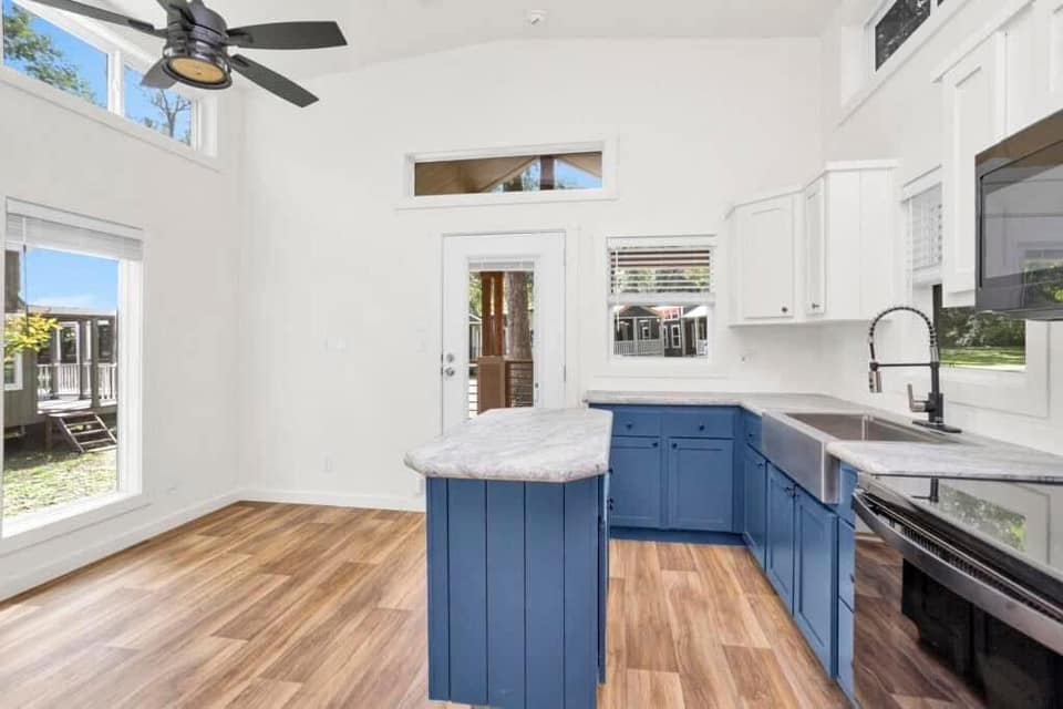Bright kitchen with wooden floors, a white ceiling fan, blue cabinets, and marble countertops. Large windows and a glass door offer abundant natural light.