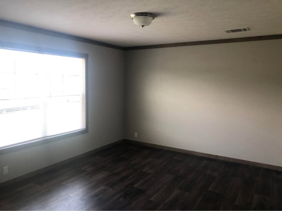 Empty room with dark wood flooring, white walls, and a large window on the left letting in natural light. Ceiling light fixture centered. Minimalist and calm atmosphere.