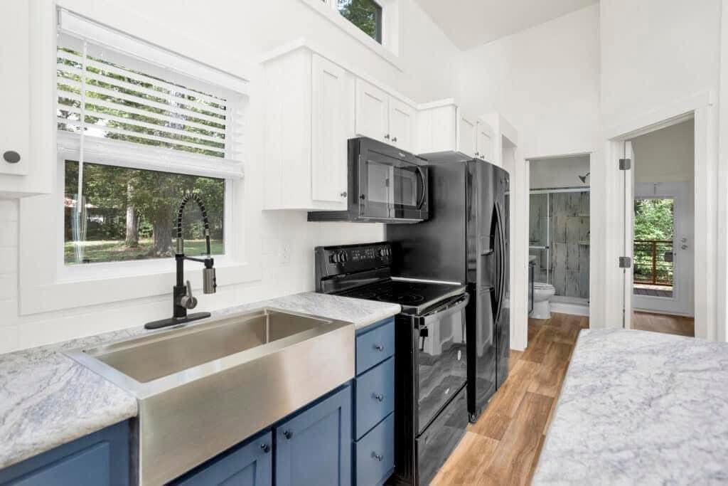 Bright kitchen with blue lower cabinets and marble countertops. Black appliances, farmhouse sink, and window with blinds enhance the modern, airy space.