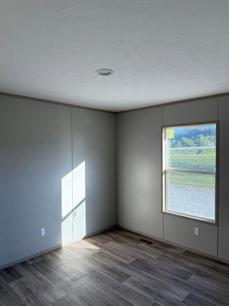 A small, empty room with gray walls and wood-like laminate flooring. Sunlight enters through a window, casting shadows. Outside, grass and gravel are visible.