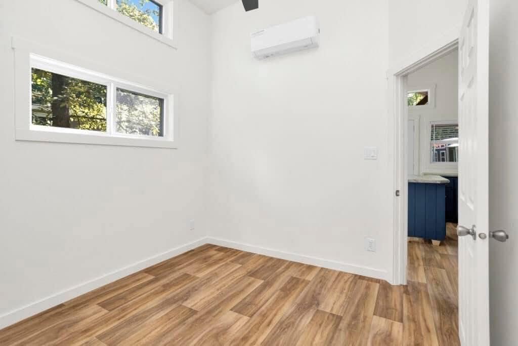 Small, empty room with white walls, light wood flooring, and two rectangular windows. Open door leads to another room with blue cabinetry. Quiet, minimalist feel.