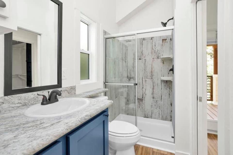A bright bathroom featuring a marble countertop with a blue cabinet, a sink, a mirror, and a shower with glass doors and rustic wall tiles. The atmosphere is fresh and modern.