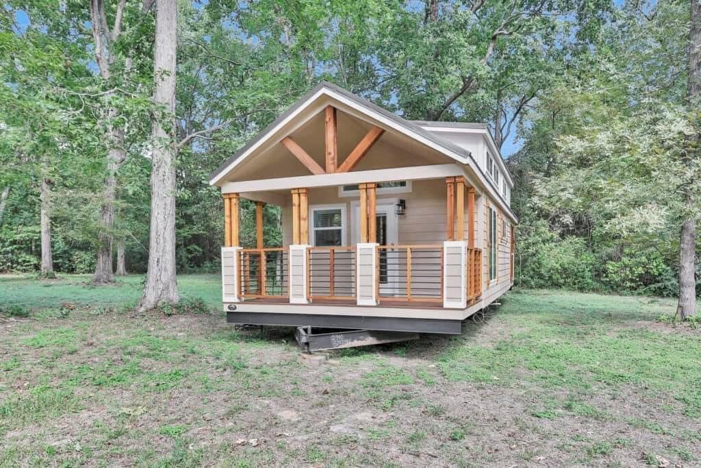A cozy tiny house with a wooden porch sits in a lush green forest. The structure is surrounded by tall trees, creating a serene, peaceful atmosphere.