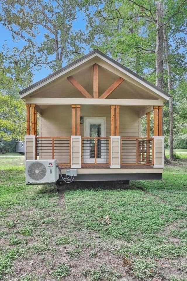 Small beige cabin with wooden beams and porch railing, set in a lush green forest. An air conditioning unit is attached on the left. Peaceful atmosphere.