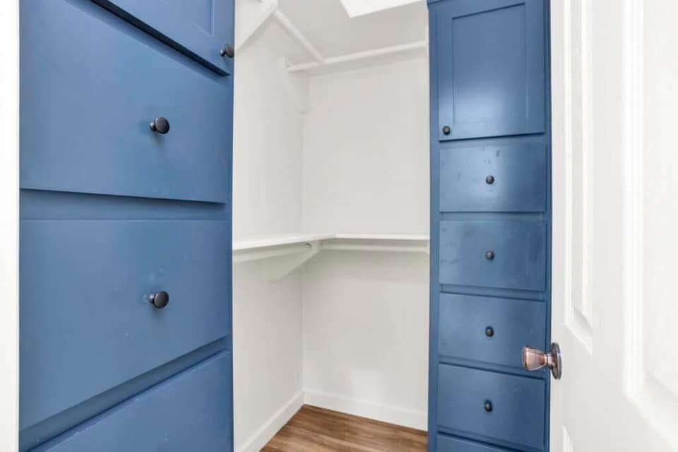 Small walk-in closet with white walls and wood flooring. Features blue cabinets with round black knobs, creating a clean and organized atmosphere.